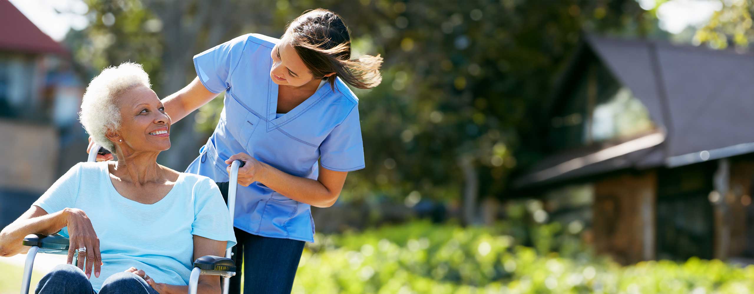 Smiling caregiver assisting an elderly woman in a wheelchair outdoors on a sunny day